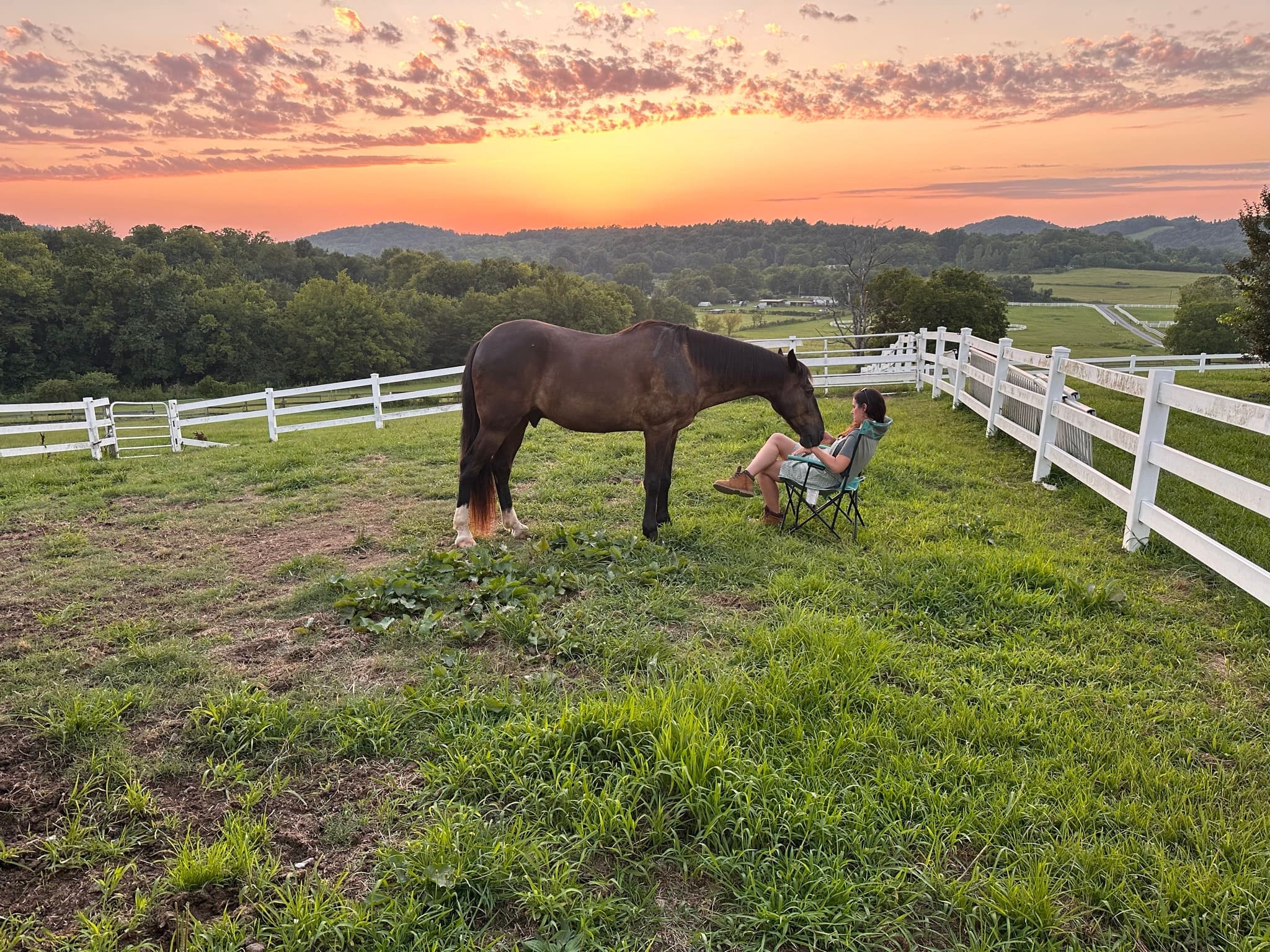 A woman sitting in a chair on the hilltop pasture at sunset, her horse grazing beside her, with rolling hills and an orange sky beyond